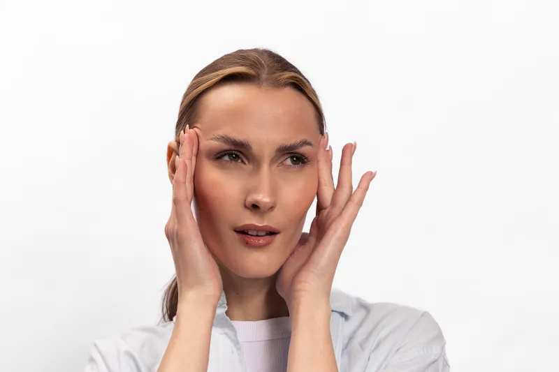 Young woman with clear skin touches her face with both hands, looking thoughtful. Isolated on white background, concept for skincare, beauty, and natural health care routine.
