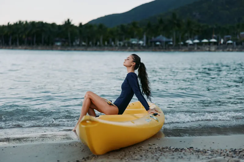 Relaxing woman sitting on yellow kayak by the sea shore wearing a dark swimsuit during calm weather.