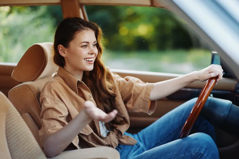 Young woman smiling while driving a vintage car, dressed in a casual shirt and jeans, enjoying a sunny day outdoors.