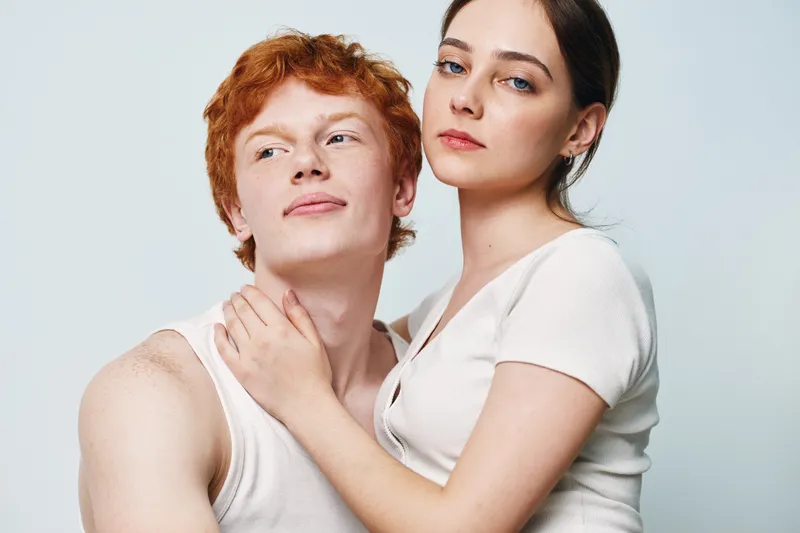 Couple posing portrait with woman embracing man tenderly, young adults in casual white tops looking confident, studio background with soft lighting.