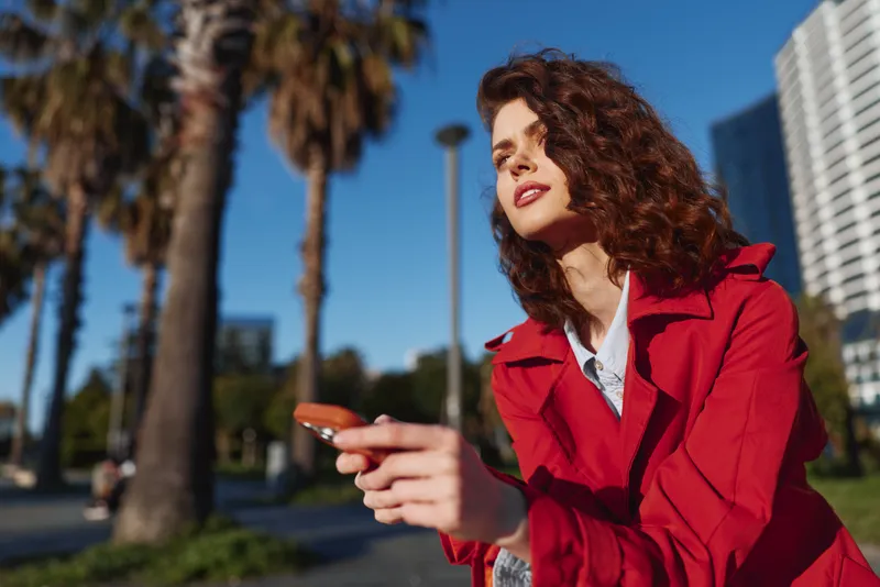 Young woman with curly hair wears a red jacket while using a smartphone outdoors on a sunny day with palm trees and urban buildings in the background.