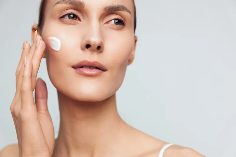 Young woman applying white face cream on cheek close-up on light background. Concept of skincare, beauty routine, moisturizing, self-care, and healthy skin with natural light.
