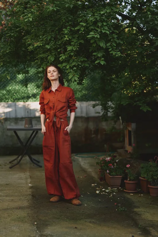 A woman in a red jumpsuit stands outdoors in a garden, a casual lifestyle portrait with confident fashion and relaxed vibe under natural light