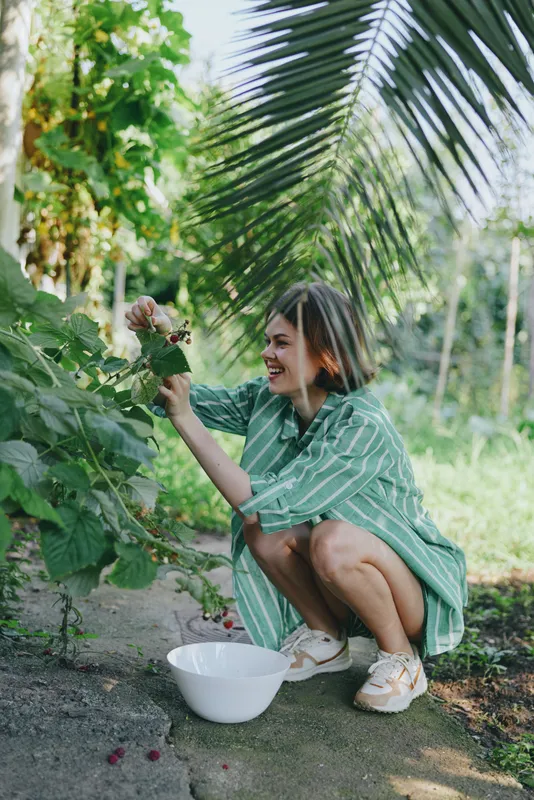 Woman crouches in a lush garden, picking berries from a bush with a white bowl nearby.