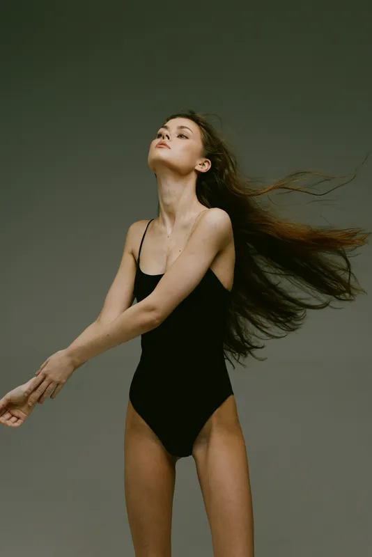 graceful woman in black bodysuit dancing with flowing hair on gray background in studio