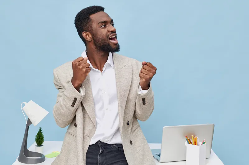 Enthusiastic young African American man in casual business attire celebrating success at workspace, fists clenched and smiling broadly, with modern office background and desk accessories, joyful mood,