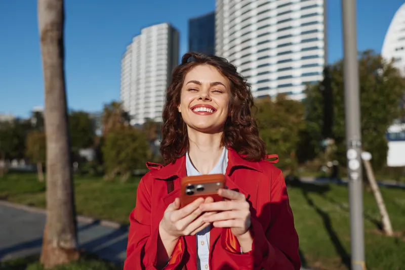 Young woman with curly hair smiles joyfully while holding a smartphone outdoors in a city park on a sunny day.