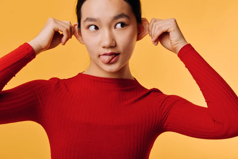 Young woman pulling ears and sticking out tongue with funny face in red sweater on orange background. Playful female portrait with expression and casual style in studio.