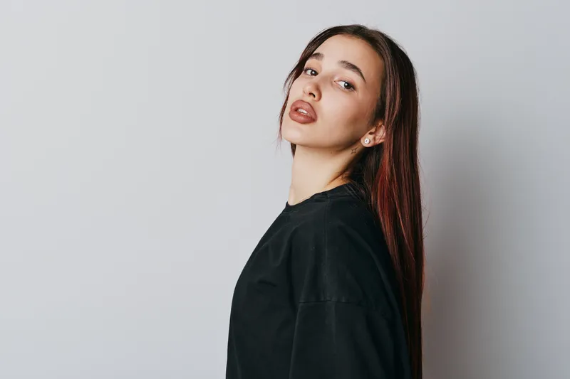 Young woman with long straight hair wearing black shirt posing against white background looking at camera with confident expression and natural makeup in studio portrait photography.