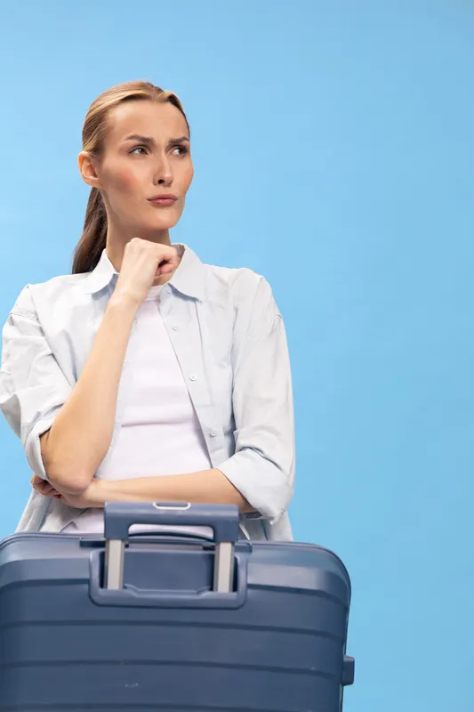 Thoughtful woman with suitcase looks away with hand on chin, wearing casual white shirt on blue background. Concept of travel planning, waiting, and decision making.