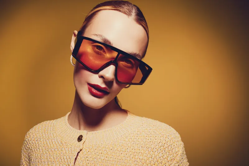Stylish woman with slicked hair wears oversized orange sunglasses and a beige knitted sweater against a warm yellow background. Concept of fashion, accessories, and modern style.