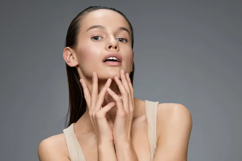 Young woman posing gracefully with soft expressions, wearing minimal makeup and a neutral outfit, highlighting beauty and confidence on a muted gray background