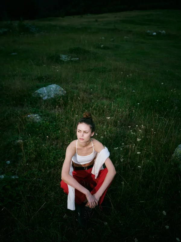 Woman sitting on grass in an open field at dusk, wearing red pants and white top with scarf, contemplative mood in a natural outdoor setting with moody lighting and soft evening atmosphere