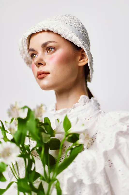 Elegant young woman in white attire holding flowers, showcasing a serene expression against a soft background, perfect for themes of nature and femininity