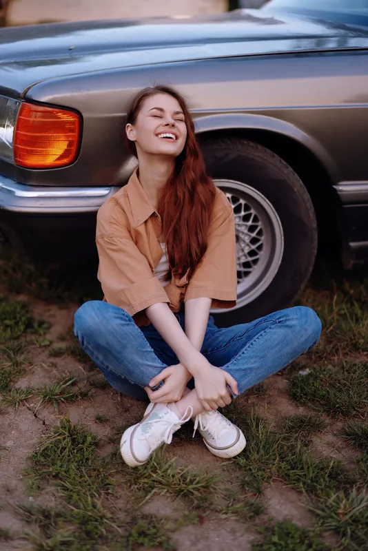 A joyful young woman in casual attire, sitting on the grass near a vintage car, exuding a relaxed vibe on a sunny day.