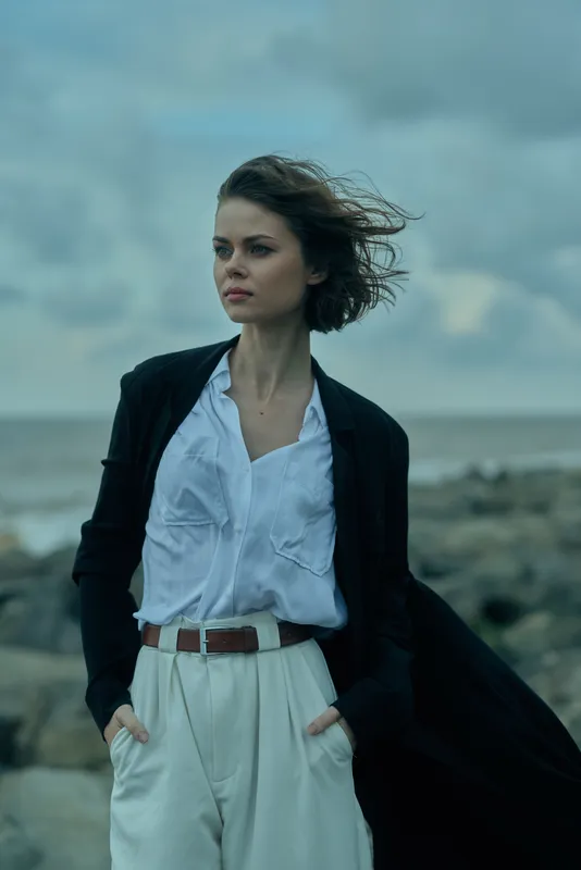 Fashion, woman and flowing coat against a windswept shoreline, white shirt tucked into light pants, coastal breeze creating movement and a chic, minimalist style suitable for editorial and lifestyle