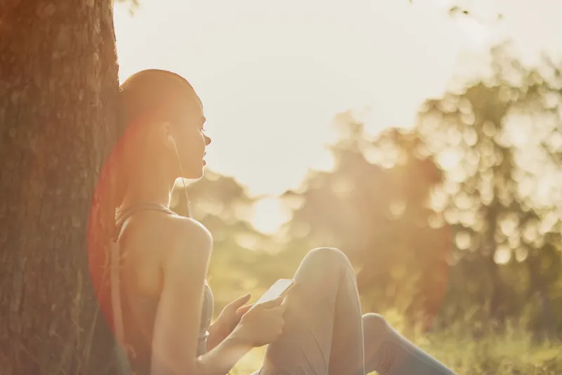Young woman listening to music while sitting under a tree, enjoying the serene golden hour light, creating a peaceful atmosphere and a sense of relaxation