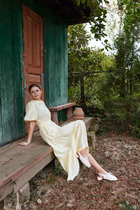 Woman wearing a pale yellow dress sits on a wooden bench beside a green cottage, relaxed pose in an outdoor garden setting with trees and earthy ground, soft sun and calm mood overall