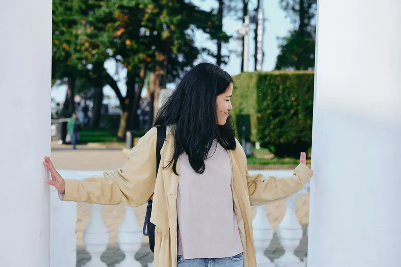 Woman in beige coat and soft sweater stands between white columns in a park, outdoor casual fashion, serene greenery and afternoon light, creating a peaceful atmosphere.