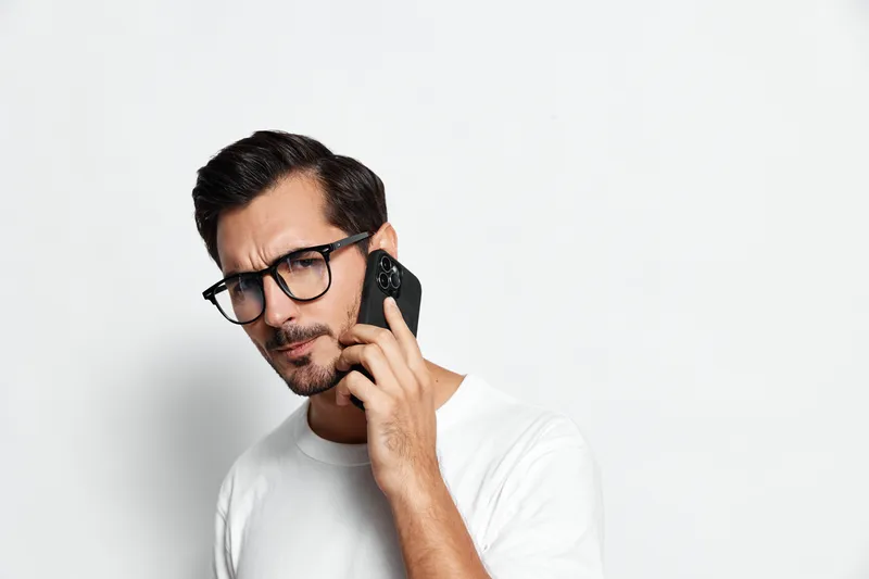 Thoughtful man in white t-shirt wearing glasses talking on smartphone, looking serious while standing against plain light background. Communication lifestyle concept.