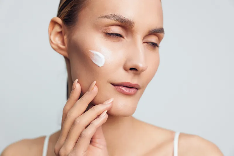Young woman with closed eyes applying white face cream on cheek, demonstrating skincare routine and self-care, soft natural lighting, neutral background, healthy skin concept.
