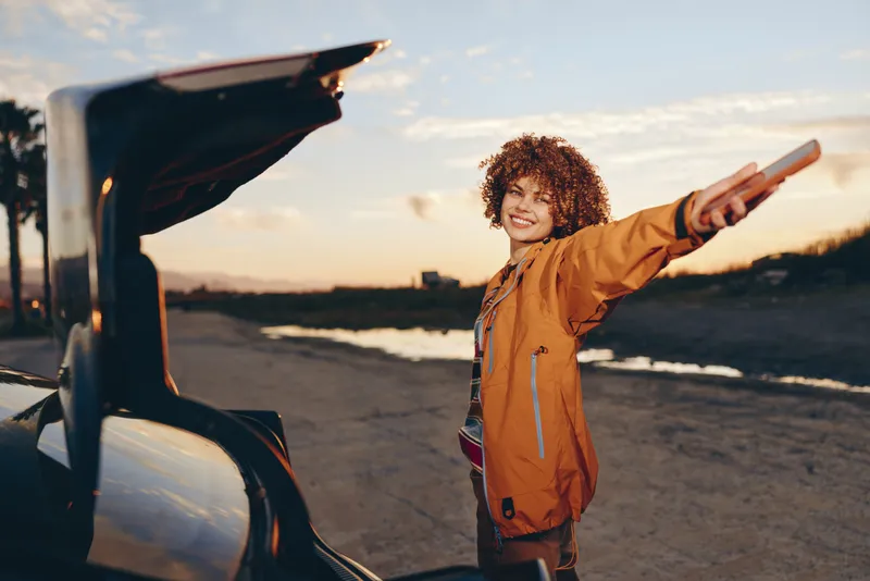 Woman lifestyle outdoors, smiling in warm sunset, curly hair, casual jacket, standing beside car on quiet road, relaxed posture, coastal evening background, natural colors, sunlit breeze