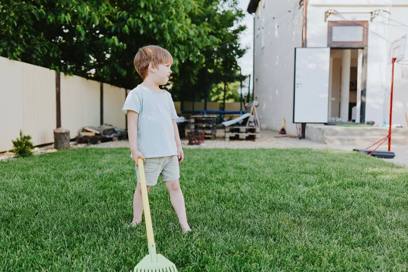 Playful boy enjoying outdoor activities, standing on green grass with a gardening rake, bright sunny day in the backyard, reflecting joy and innocence.