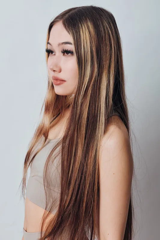 Young woman with long straight hair and natural makeup posing in profile on white background. Beauty model wearing neutral top with calm expression and soft lighting.