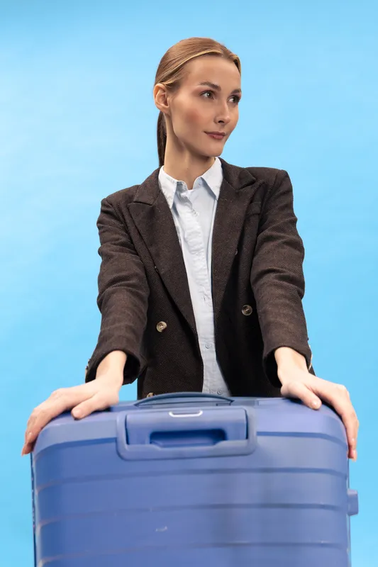 Young woman in brown blazer stands behind blue suitcase with hands on handle, looking to side. Concept for travel, business trip, and modern lifestyle on blue background.