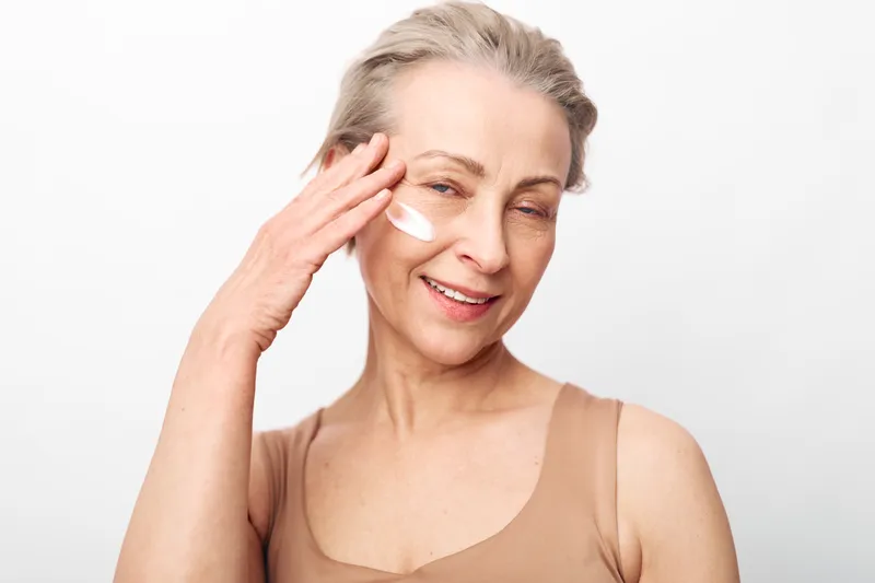 Smiling mature woman with light skin applies moisturizer cream on face, enjoying self-care and skincare routine. Studio portrait with white background and natural beauty concept.