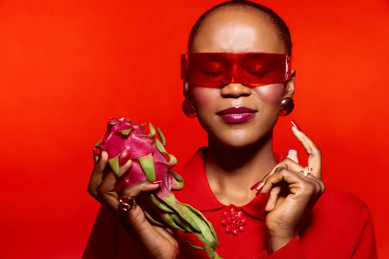 Confident African woman with dark skin holds a dragon fruit, wearing red futuristic glasses and matching red outfit against a vibrant red background. Concept of fashion and fruit.