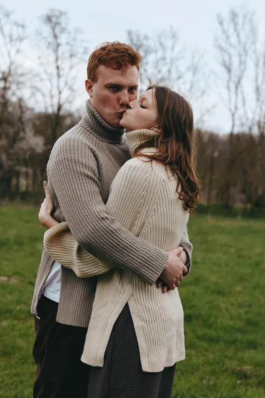 Young couple embraces and shares a tender kiss outdoors in cozy knit sweaters during autumn, expressing warmth and affection in a natural lifestyle setting with soft daylight.