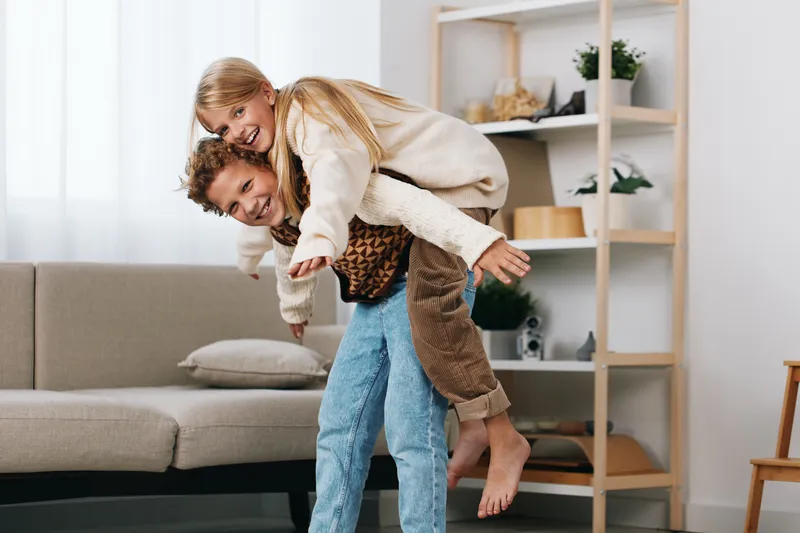 Playful couple shares a joyful piggyback moment indoors, smiling as the partner carries the other on the back in a cozy living room with soft light, creating a candid, energetic vibe.