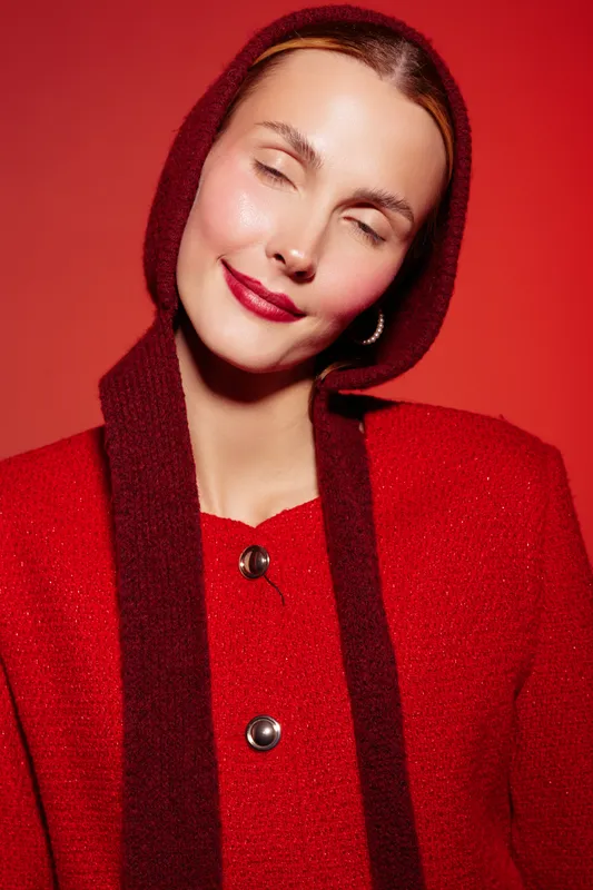 Young woman in red sweater and hood smiles softly with closed eyes against red background. Studio portrait with warm lighting and cozy fashion concept for autumn or winter style.