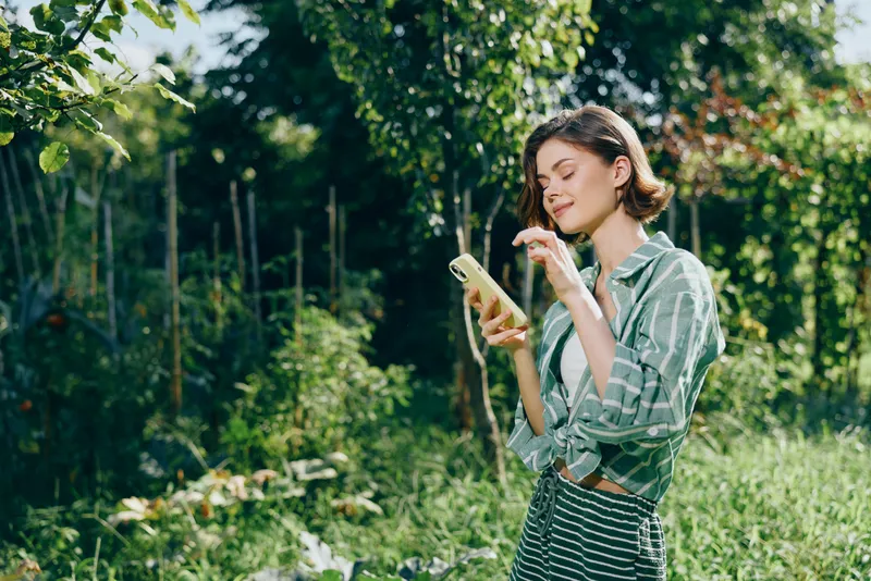 Young woman stands in a bright garden, looking at her smartphone and enjoying the warm outdoor light.