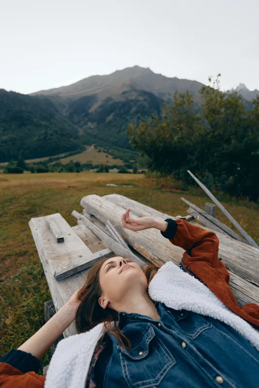 Woman lying on wooden bench in mountains, relaxation in nature and outdoors meadow scenery, casual denim jacket and phone on table, peaceful portrait of leisure escape.