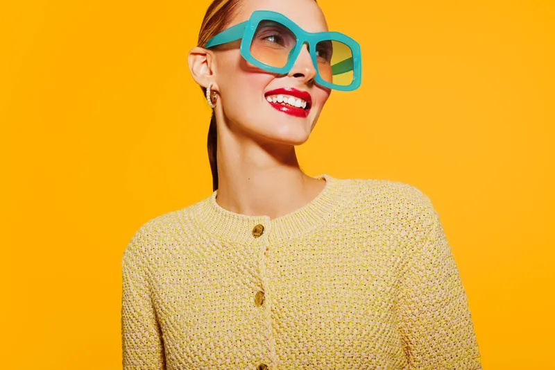 Stylish woman with bright red lipstick and large turquoise sunglasses smiles confidently against vibrant yellow background. Fashion concept with bold colors and modern accessories.