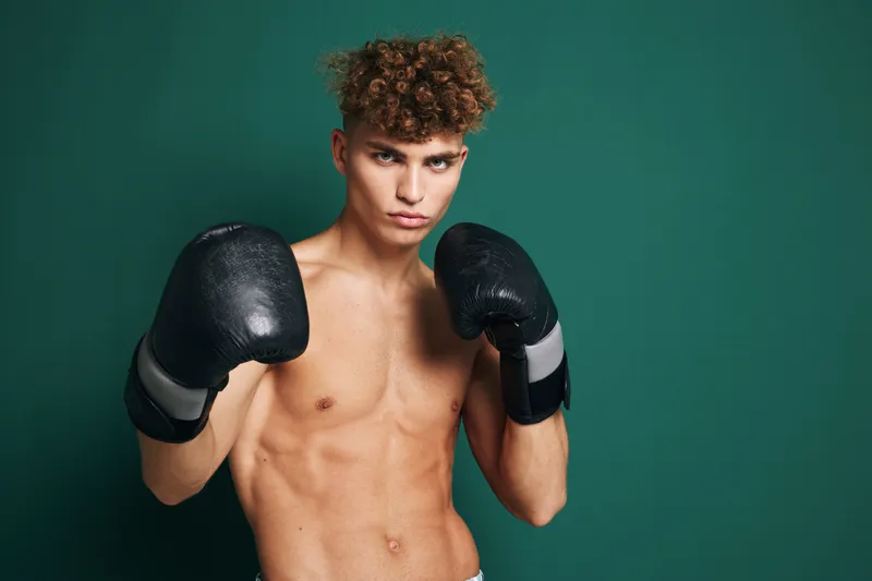 Young male boxer with curly hair wearing black boxing gloves in fighting stance against green background. Athletic shirtless man training for boxing or martial arts.