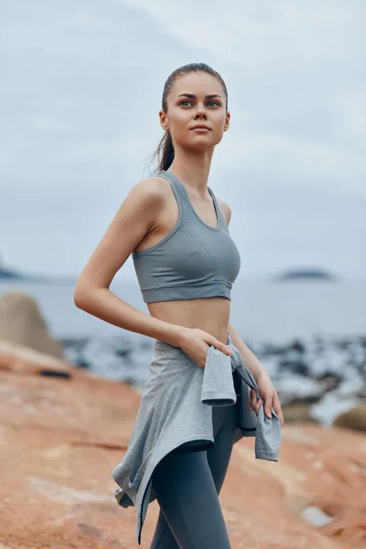 Woman in athletic wear with a confident expression, standing by the ocean on a rocky beach, showcasing an active lifestyle and fitness concept in a seasonal, natural setting