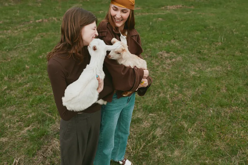 Two women outdoors cuddle white goat kids, smiling and sharing a moment of rural life, with natural daylight on a grassy field, casual jackets, warm mood, friendly vibe, and gentle animal interaction.
