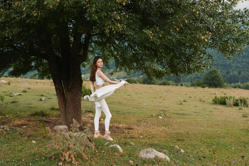 Young woman in white activewear spins joyfully near a large tree in a grassy field surrounded by hills under a cloudy sky.