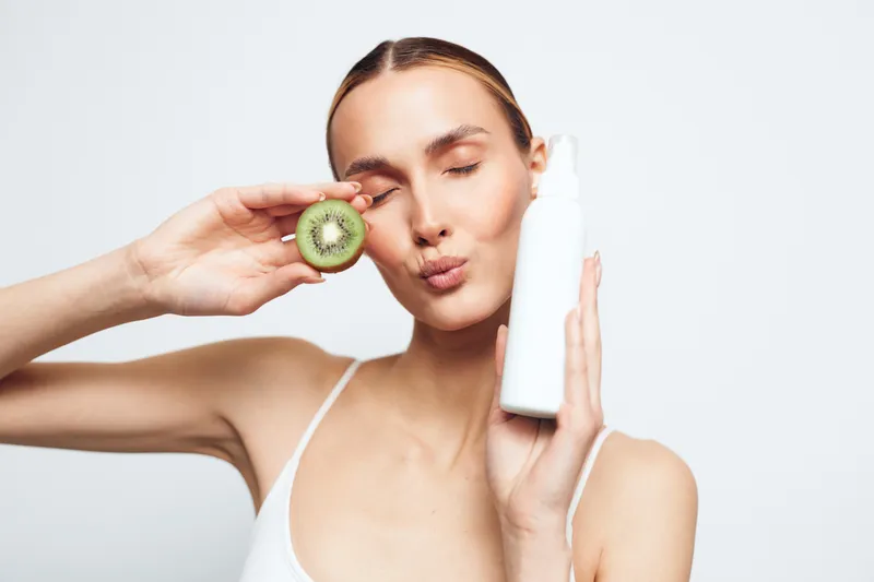 Young woman holding a white cosmetic bottle and a sliced kiwi near her face with closed eyes. Concept of natural skincare, beauty, and healthy lifestyle on white background.