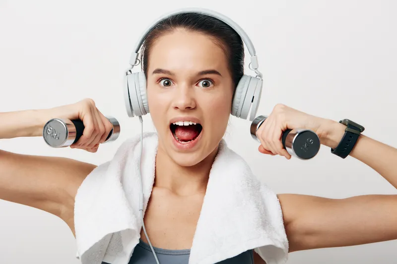 Fit woman fitness sport scene: young female exercising with small dumbbells and over ear headphones, white towel draped over shoulders, dynamic energetic expression, studio white background closeup