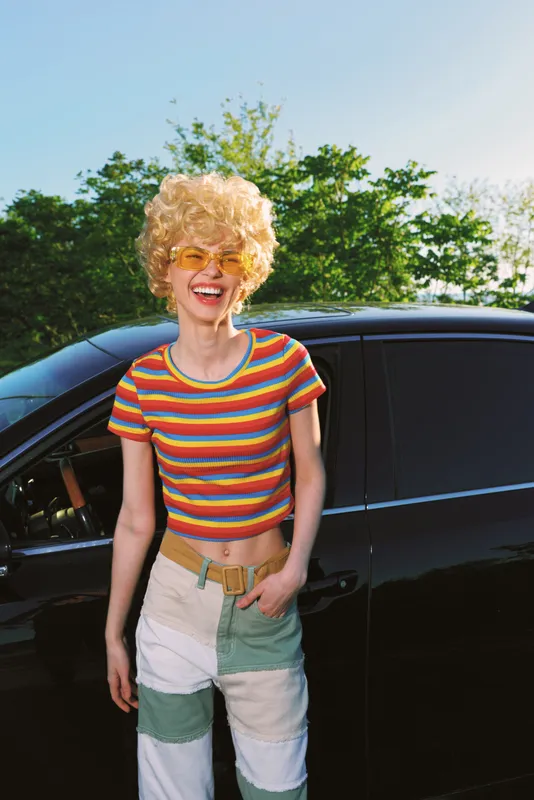 Young woman with curly blonde hair wears a rainbow striped crop top and light patchwork pants, smiling beside a dark car on a sunny day.