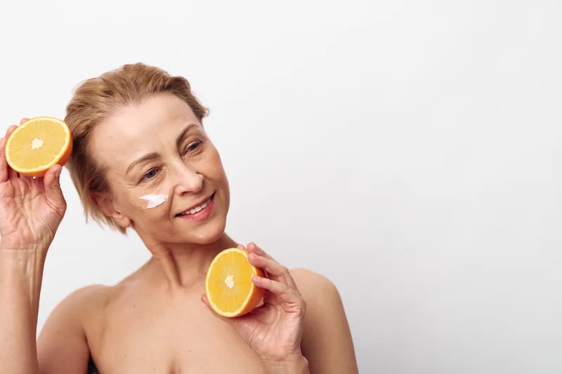 Smiling mature woman with short hair holds halved orange near face with facial cream patches on cheek, isolated on white background. Concept of natural skincare, vitamin c, and healthy beauty routine.
