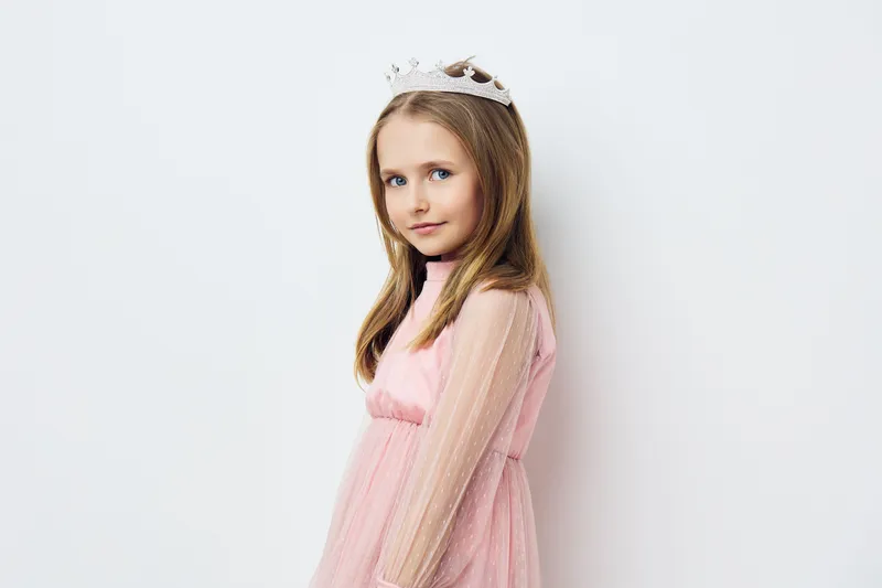 Young girl in a pink dress with a crown, looking confidently at the camera, set against a light background, embodying innocence and joy