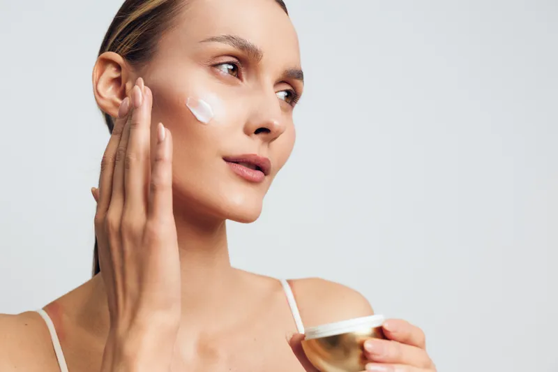 Young woman applying white face cream on her cheek with fingers. Close-up portrait on light background. Concept of skincare, beauty routine, and moisturizing. Soft natural lighting.