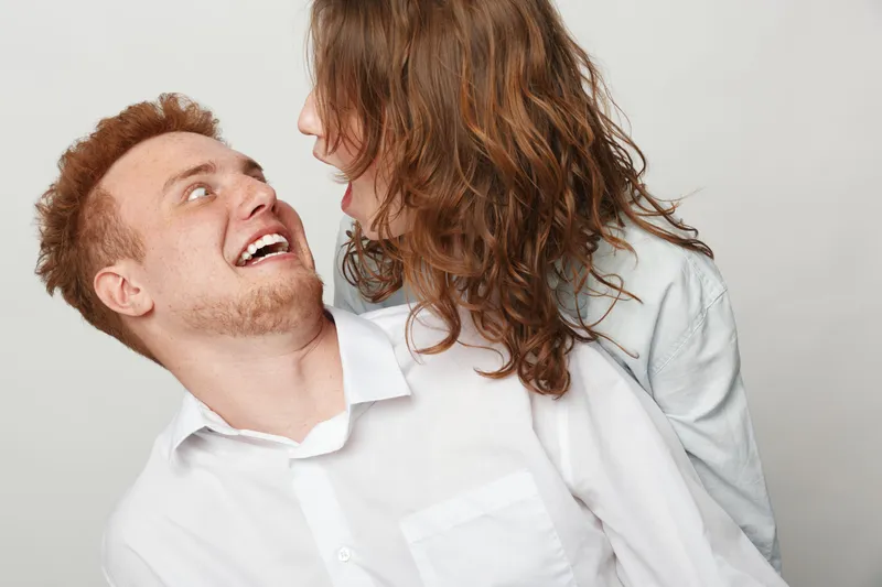 Happy young couple in white shirts looking at each other with joy and laughter. Close-up portrait on light background. Concept of love, relationship, and positive emotions.