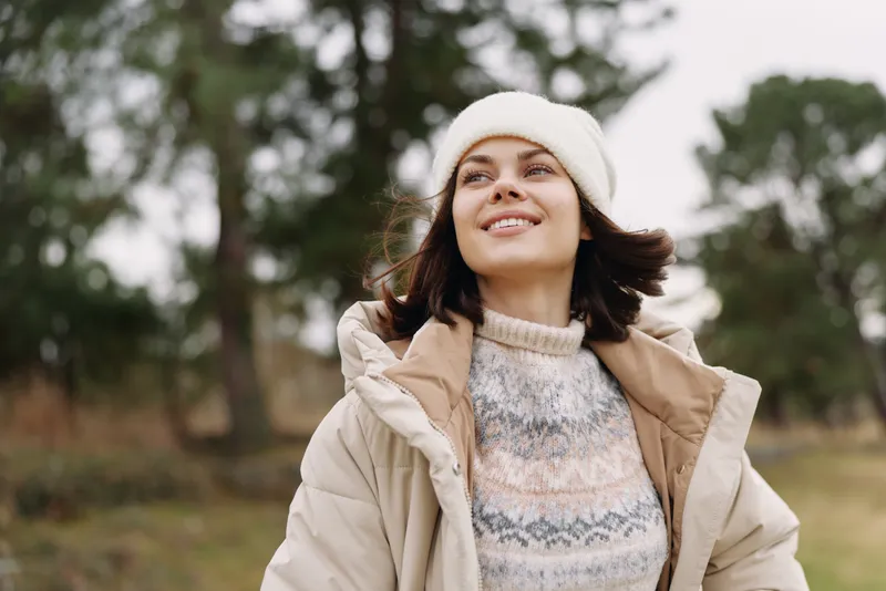 Woman in an outdoor setting wearing a warm coat and cozy sweater, smiling under a knit beanie on a windy autumn day, surrounded by trees and open field