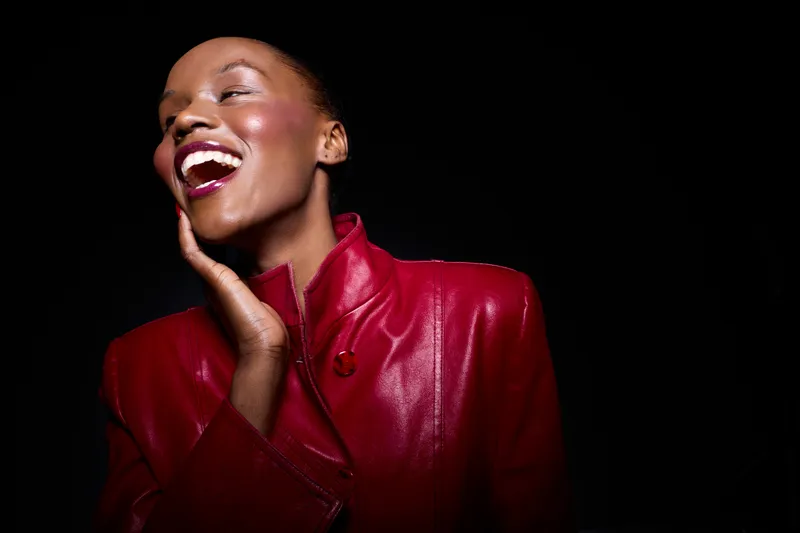 Happy dark skinned African woman laughing with closed eyes and hand on face wearing shiny red leather jacket. Studio portrait on black background. Concept of joy, fashion, and beauty.